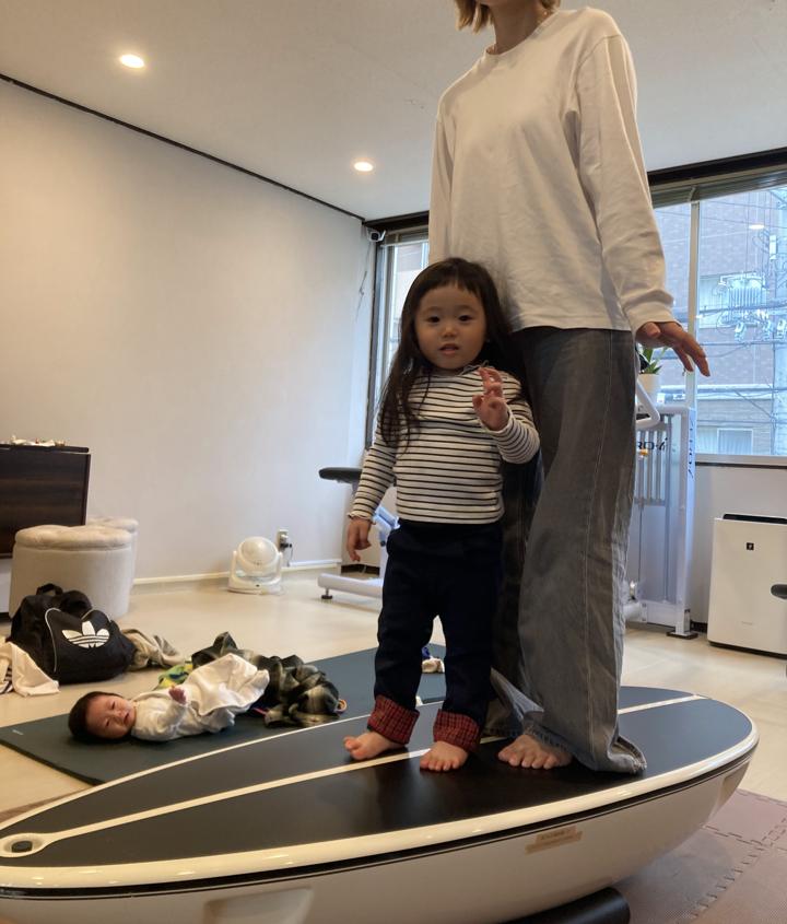 お母さんと小さな女の子が一緒にサーフトレーナーに乗り、うしろのマットでは赤ちゃんが眠っている様子