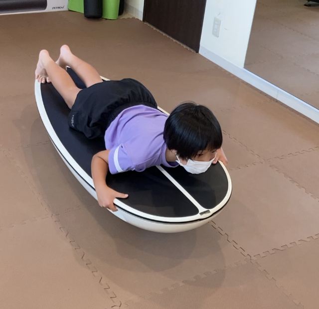 紫色のTシャツの女の子が、サーフトレーナーの上にうつぶせになってバランスをとる様子